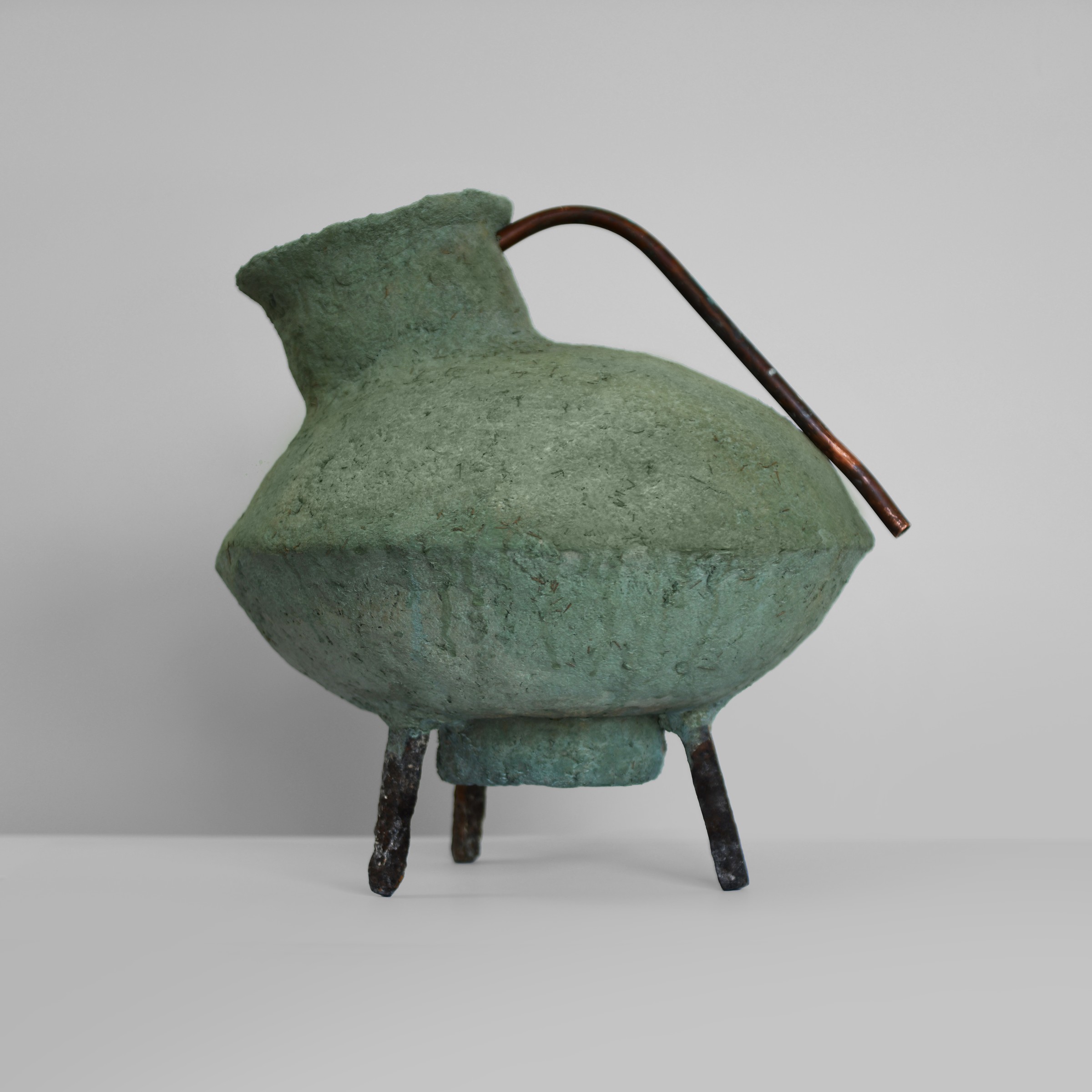 A green clay-paper vessel with a copper handle and three dark metal legs.