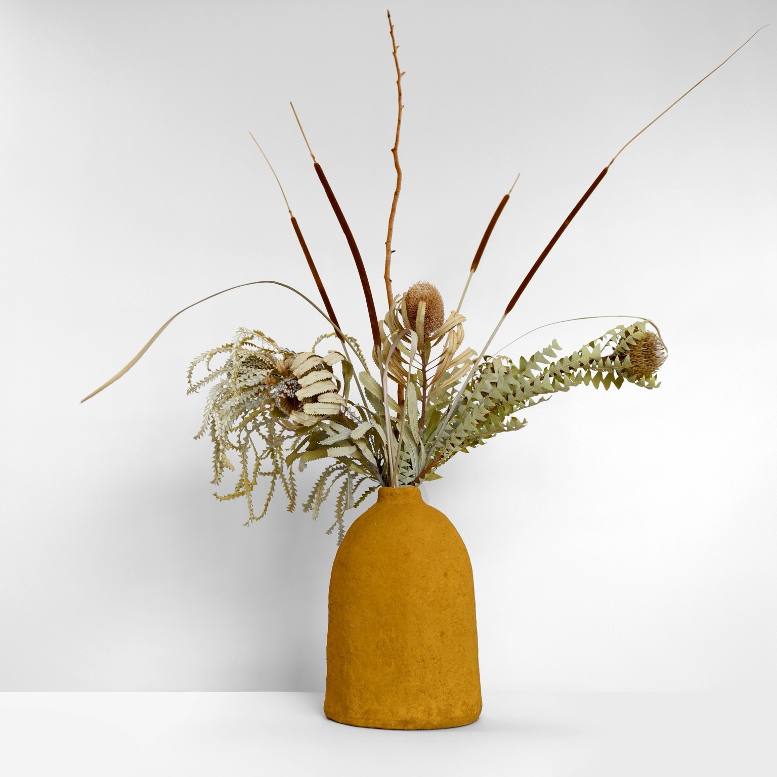 A tall ochre clay-paper form photographed with dried foliage and fine upright stems.